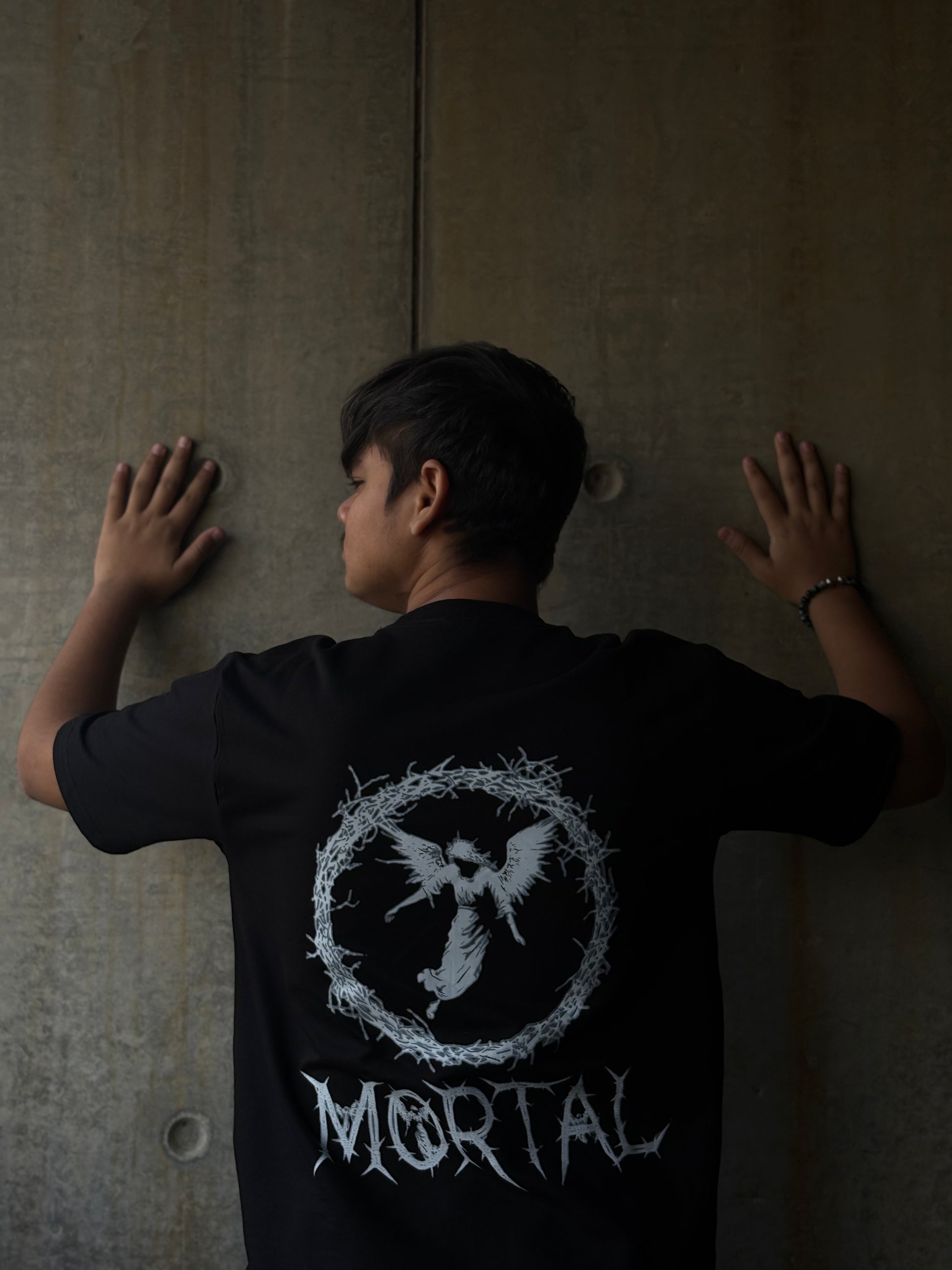 Person wearing a black t-shirt with a logo and text, standing against a plain wall.