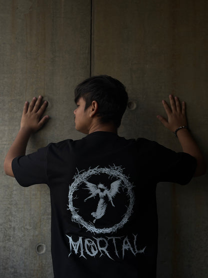 Person wearing a black t-shirt with a logo and text, standing against a plain wall.